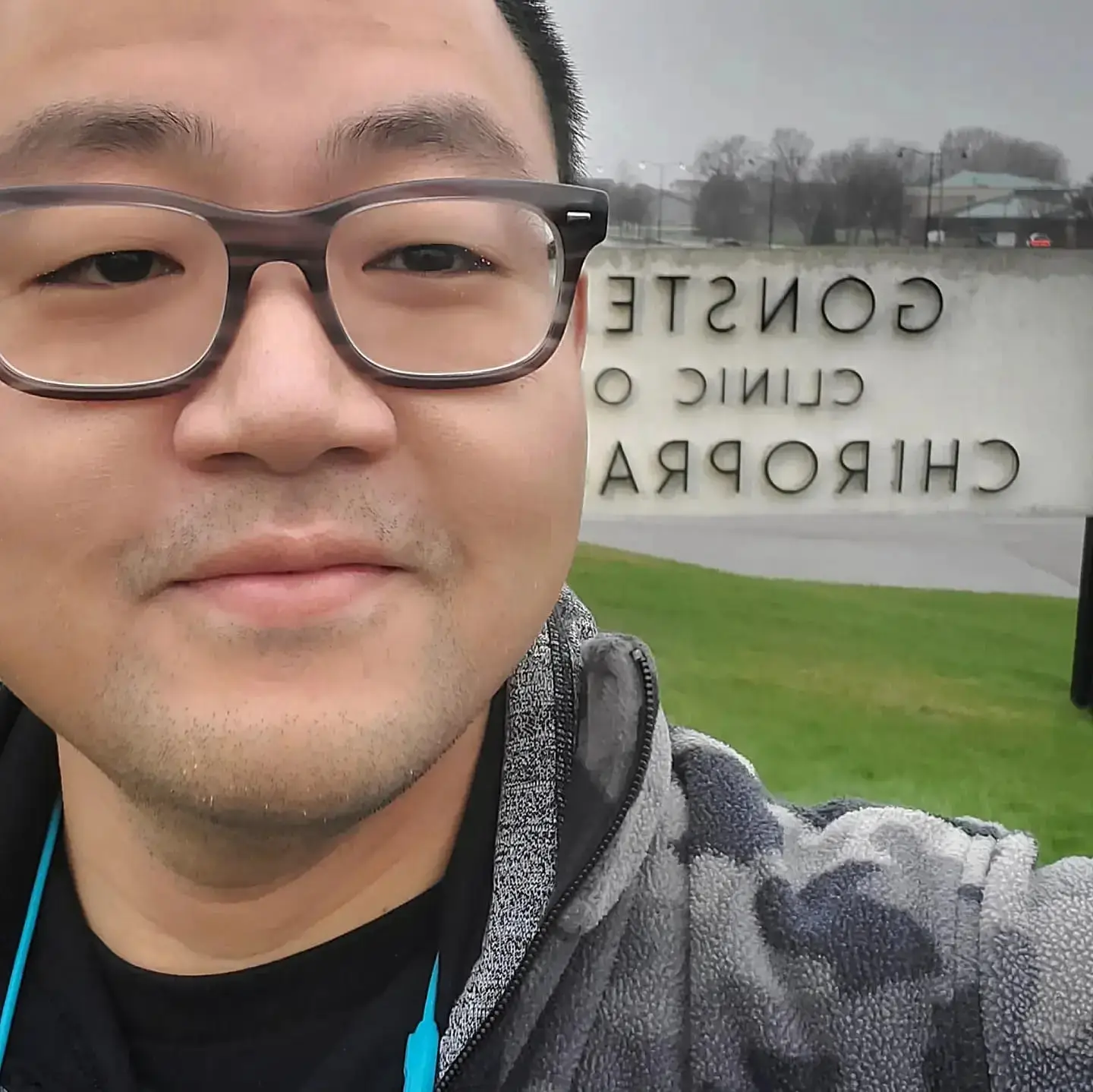 A smiling individual wearing glasses and a patterned jacket, standing outdoors in front of a sign that reads "Gonstead Clinic of Chiropractic" in reverse reflection.