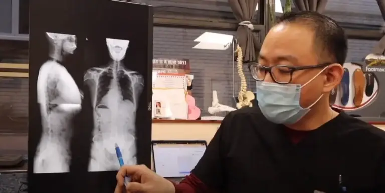 A chiropractor wearing a mask points to an X-ray image, explaining hip or spinal issues to a patient.